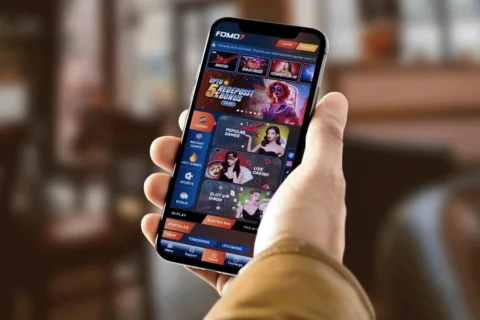 Person holding a smartphone showing the FOMO7 India app home screen with live casino and games.